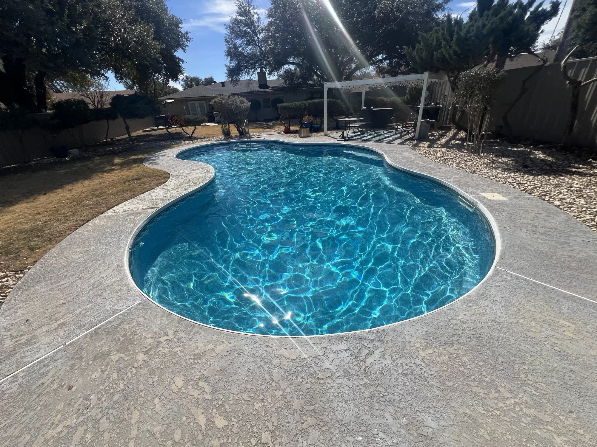 Blue freeform backyard pool beside a concrete deck and white pergola