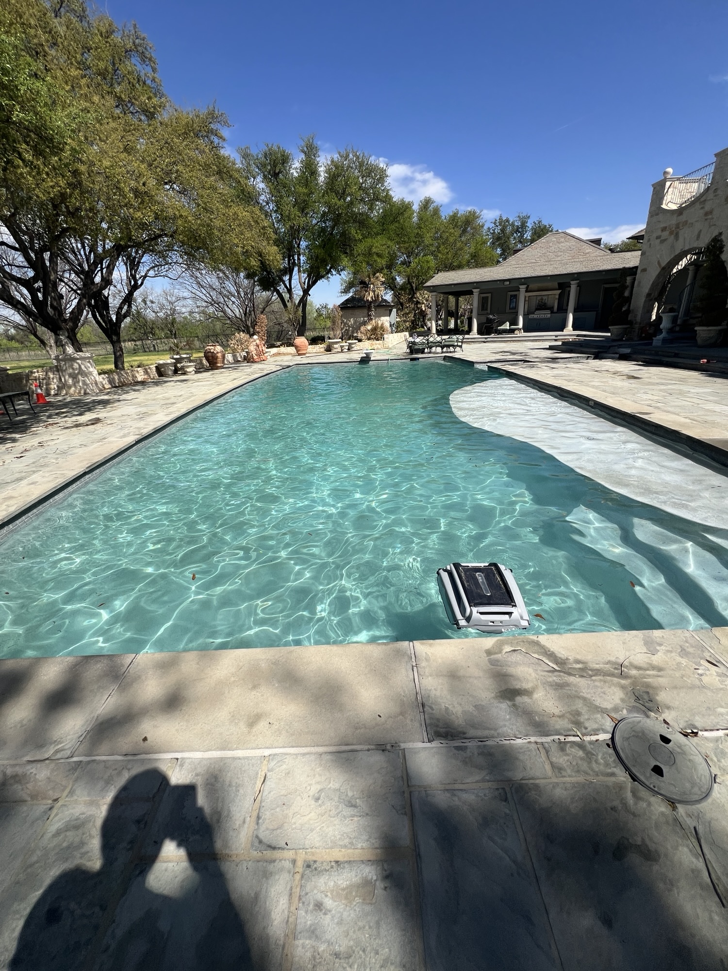 Clear swimming pool with a curved shallow shelf, stone deck and a pool-cleaning device near the foreground.