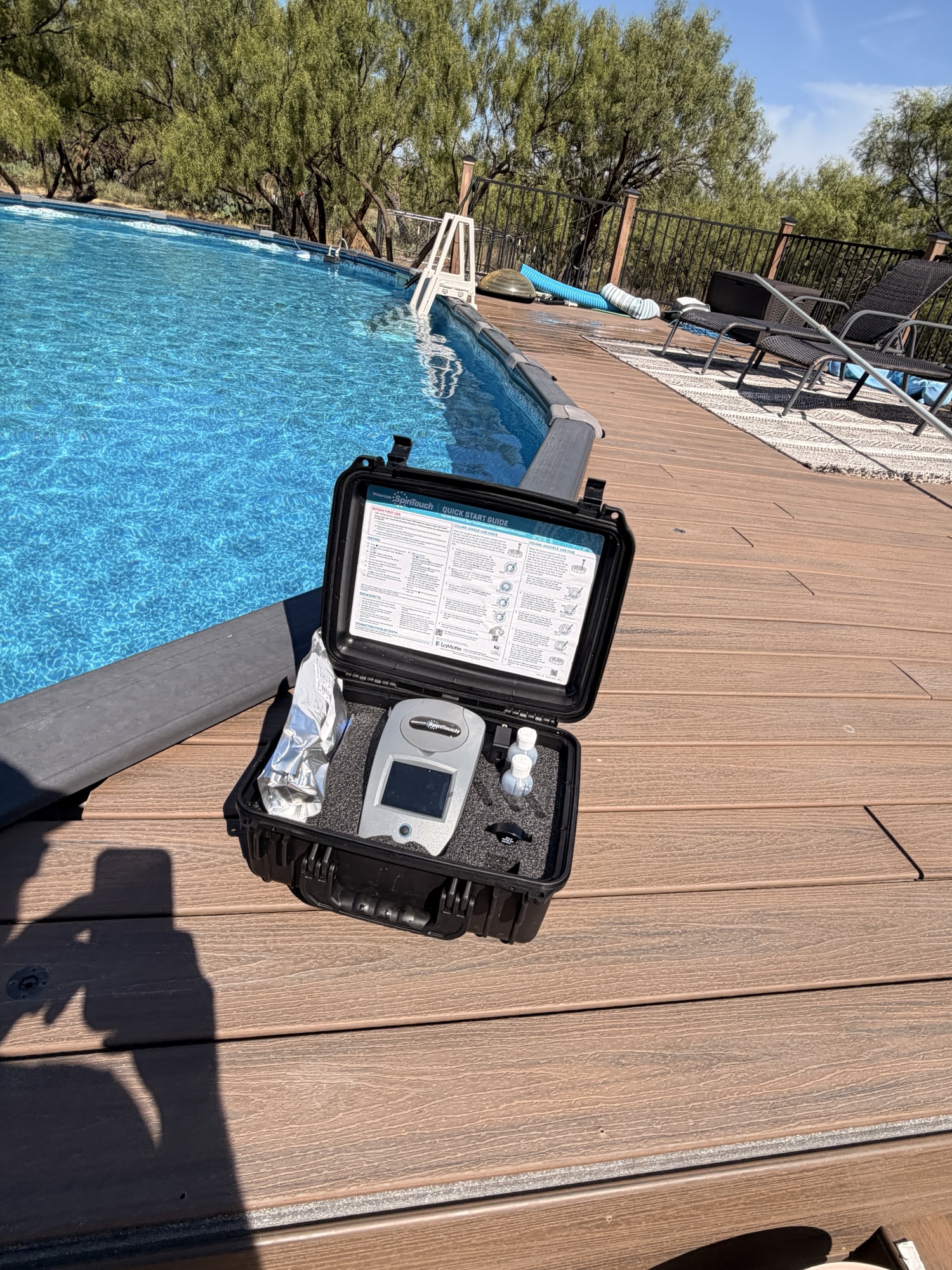 SpinTouch digital water tester in an open black case on a wooden deck beside a blue above-ground pool.